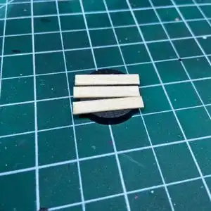 Three one inch bass wood strips glued to the top of a 10mm Warhammer base on top of a hobby cutting mat