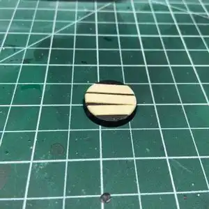 Three trimmed down one inch bass wood strips glued to the top of a 10mm Warhammer base on top of a hobby cutting mat